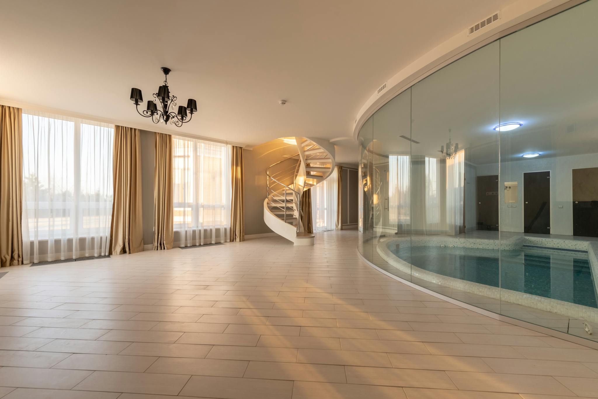 Swimming pool with clear water behind glass wall placed in modern hall with stairway and windows with curtains in contemporary house